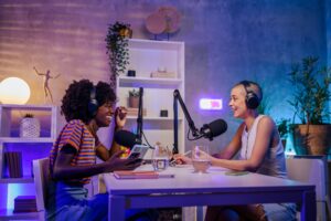 Beginners guide to Podcasts with Two cheerful podcasters are sitting in a little home podcast studio.